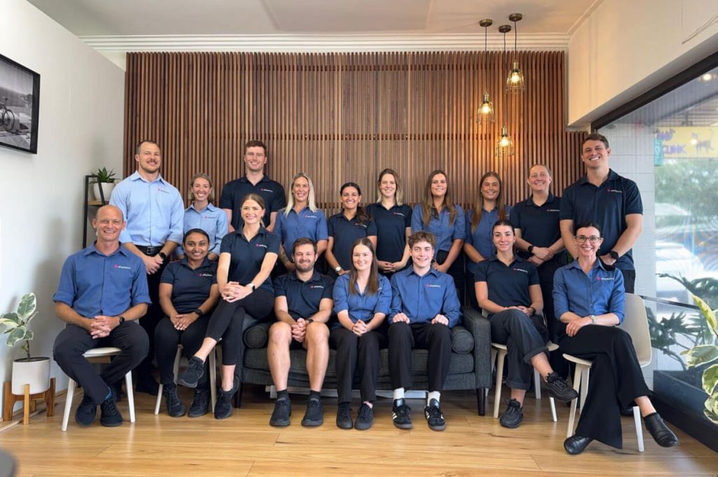A group of 18 people, wearing matching blue uniforms, pose and smile for a group photo indoors. They are seated and standing in two rows, with a modern, wooden-panel background behind them. — Physio Club